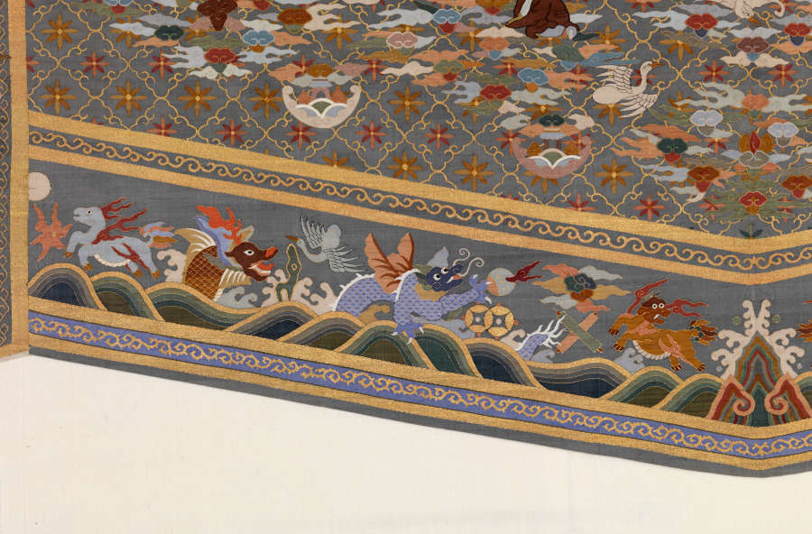 Left border detail of the triangular tail of the gray-blue robes back featuring illustrations of wavy landscape, dragons, birds, and animals encased by blue and gold patterned stripes.