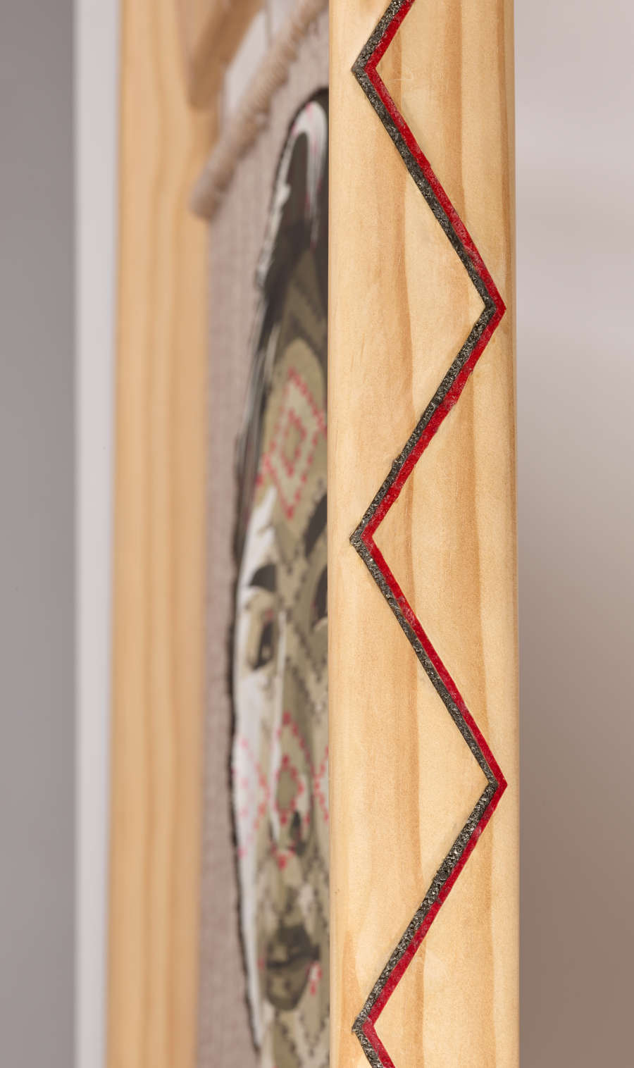Close-up of a wooden frame's corner,  which is detailed with a vertical red and black zigzag design. The wooden frame holds a natural-toned woven tapestry of a figure’s face.  