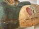 An angled view of a painted coffin with a blue-black headdress and a red disk visible on the shoulder, with signs of age and wear.