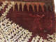 Closeup of a deep red woven fabric with geometric embroidery depicting a triangle border and a humanoid figure in red. Along the bottom are cascading reflective sequins.