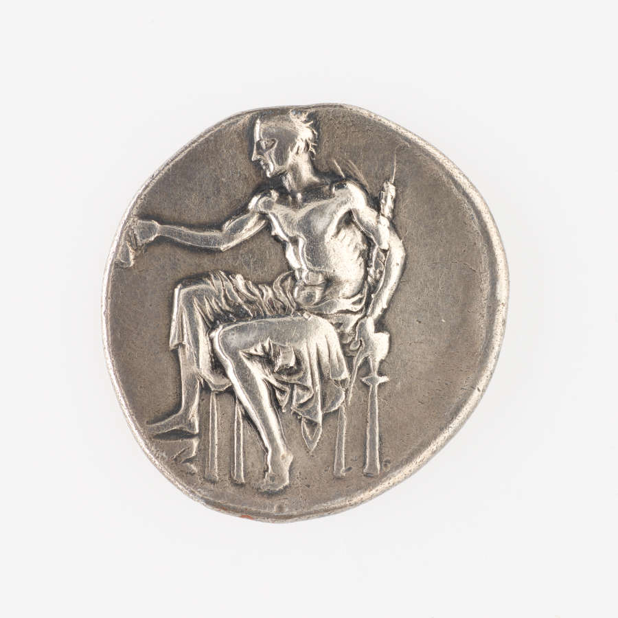 One side of an irregular round silver coin, embossed with an image of a seated man holding his hand out whilst grasping an implement in the other.