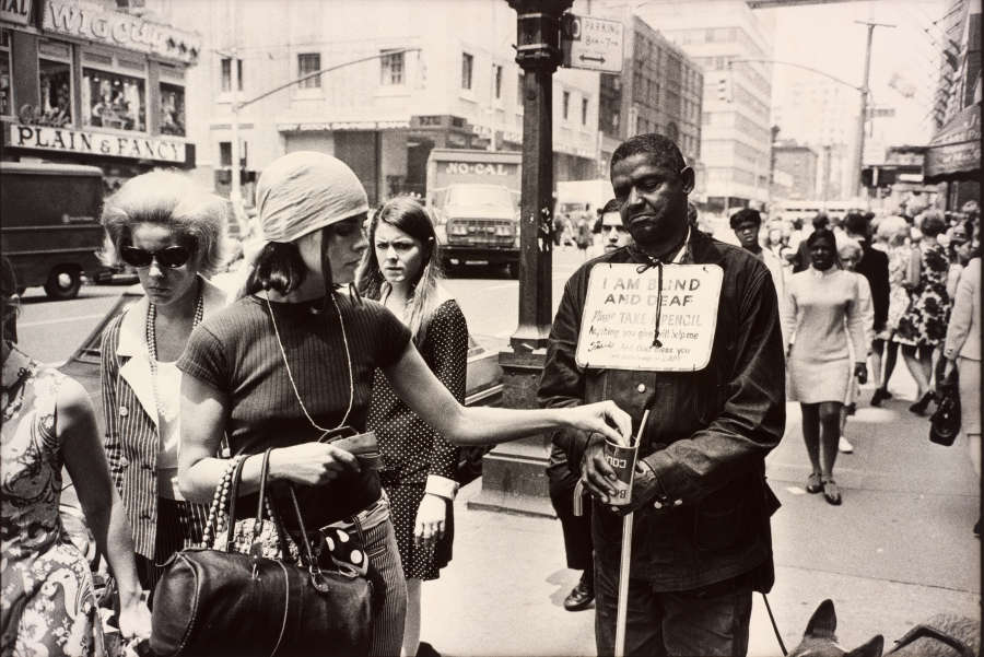 A grayscale photograph of a light-skinned person putting change into the cup of a dark-skinned person, who wears a sign around their neck that reads “I AM BLIND AND DEAF.”