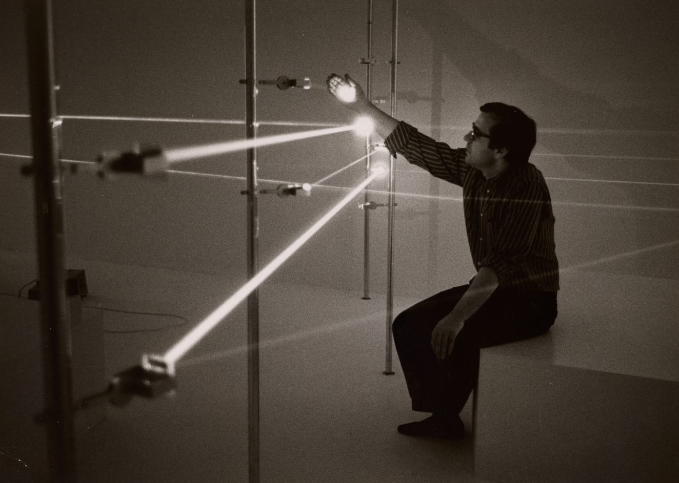 A person wearing glasses sits on a white bench in a dimly lit space, reaching out to touch a bright white laser beam as other beams intersect, criss-crossing the room. 