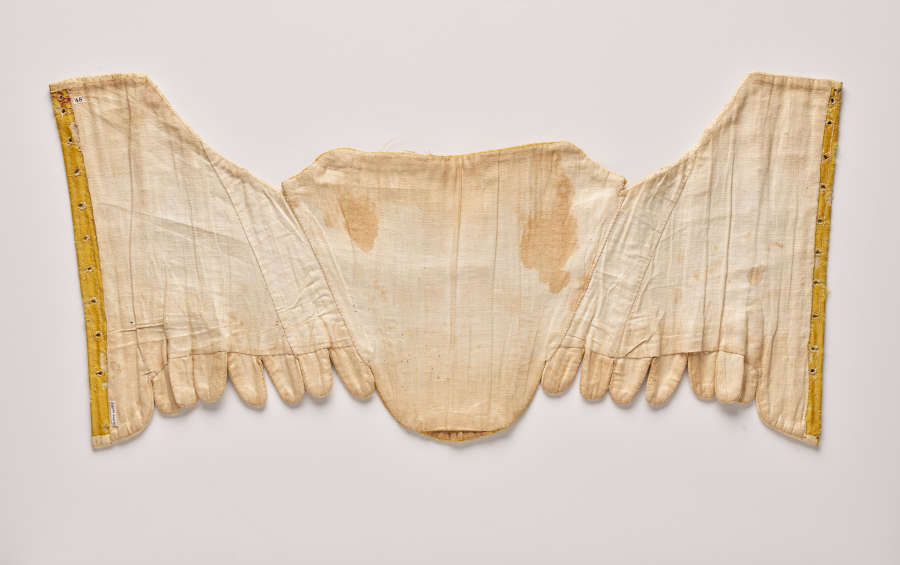  Photo of an intricate, symmetrical off-white sewn form with gold-colored edges, lying flat. Stitching is visible between panels. Two tan-colored stains appear at the top of the center panel.