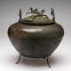 A brown-green patinated metal bowl with decorative engravings on its onion-shaped body, sitting on a three-legged stand. Metal-cast winged horses dance along the rim of the bowl’s domed lid.