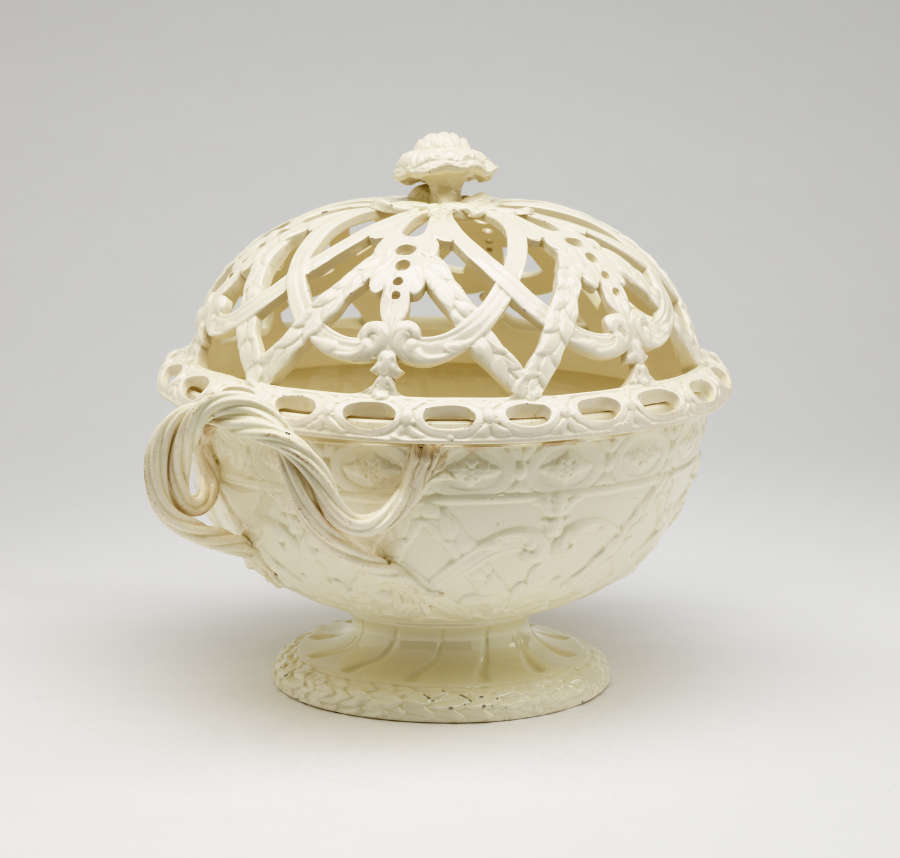 A cream-colored basket with sculptural woven handles, sculpted foot, and a lid with cutout designs.