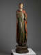 A tall, weathered wooden statue of a robed figure with a serene expression, draped in green and red garments, standing upon an octagonal base.