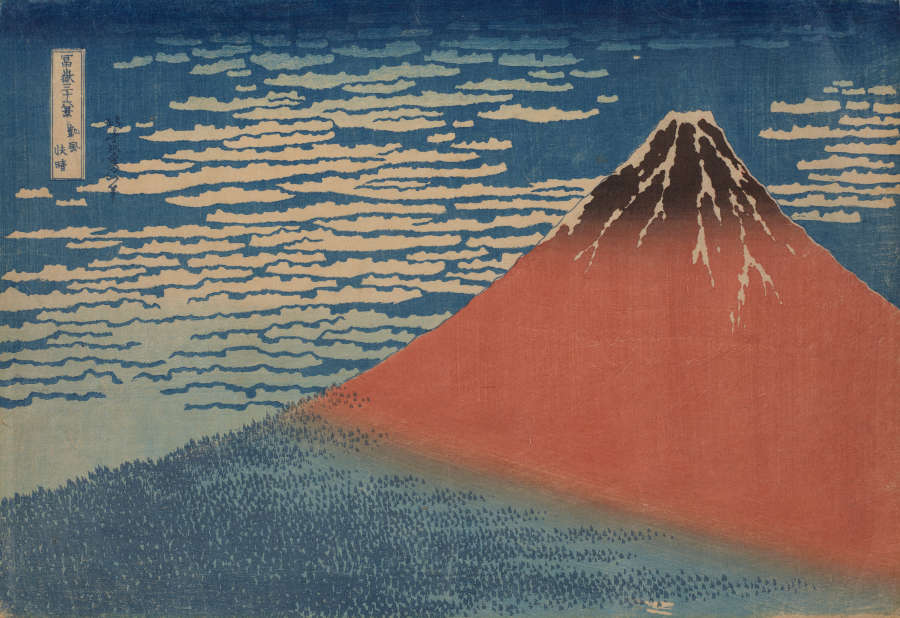 A woodblock print of a partially sunlit mountain against a cloudy blue sky. The mountain's blue shadowed base is forested whilst the red sunlit tip of the mountain is snowy.