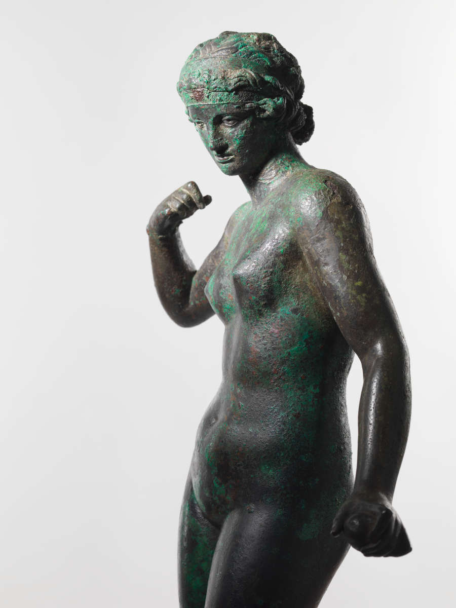 Cropped image of a gray metal sculpture with a green patina of a standing crowned nude woman, one arm down, grasping an object, and the other curled towards her shoulder.