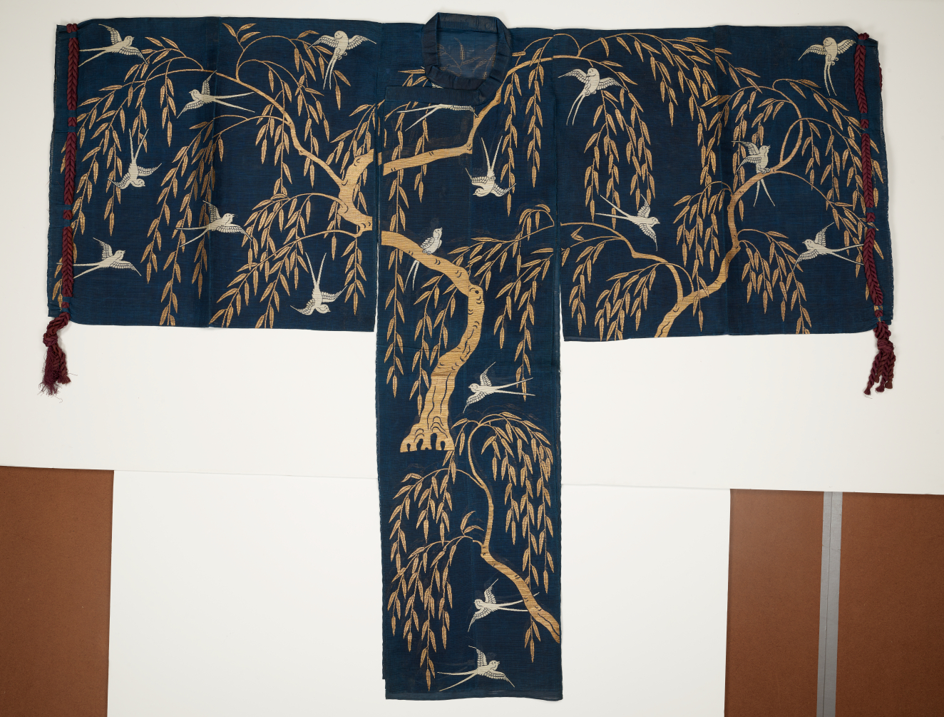 A dark blue kimono adorned with gold willow trees and white birds, displayed flat against a white background with red tassels on the sleeves. 