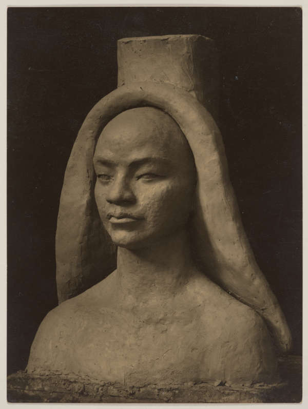 Photograph depicting the bust of a figure with a tall round headpiece, in a three quarter view. The face looks serene and the clay-like material emphasizes its textured surface.