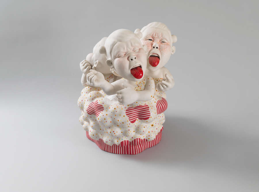Sculpture of three childlike figures with oversized heads, bright red tongue sticking out of their exaggerated mouths, emerging from a circular, cupcake-like base decorated with thin red and white stripes.