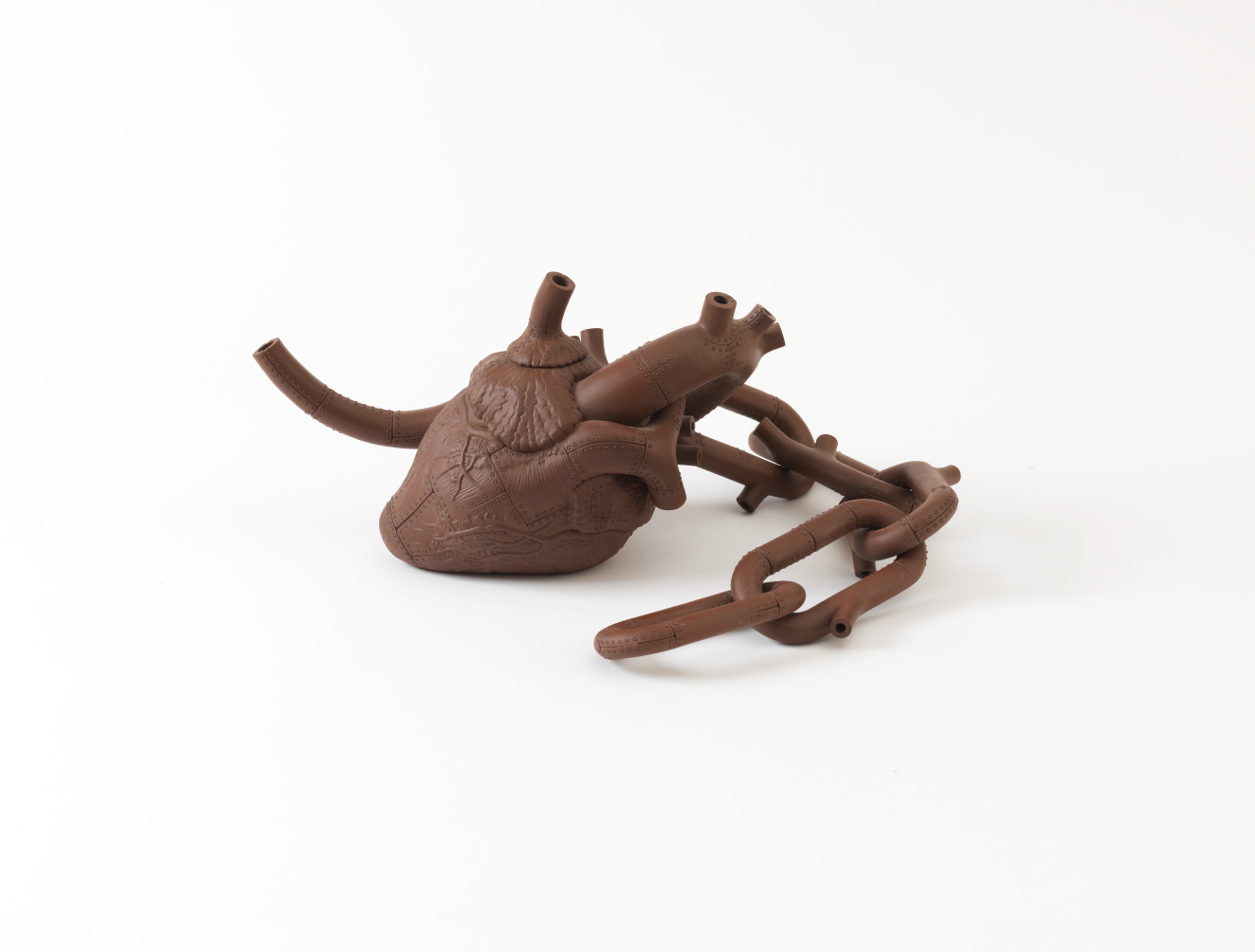 Brown ceramic teapot shaped like an anatomical human heart with fine vein-like textures on the surface and extended artery-like spouts, accompanied by a large chain link curling to the side.