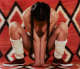 Nude person with braided hair and calf length shoes sits crouched, facing down, on a red geometric patterned backdrop. The person’s arms pressed together to cover their body.
