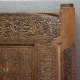 Close-up of an intricately carved wooden panel featuring ornate calligraphic inscriptions, framed by decorative floral patterns and geometric designs, with a broken section along the right corner. 