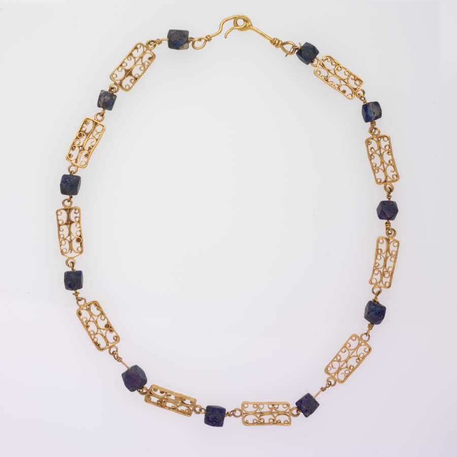 Necklace with alternating rectangular gold filigree ornaments and dark square beads, with a hook and loop fastener. The filigree grids divide each rectangle into six parts.