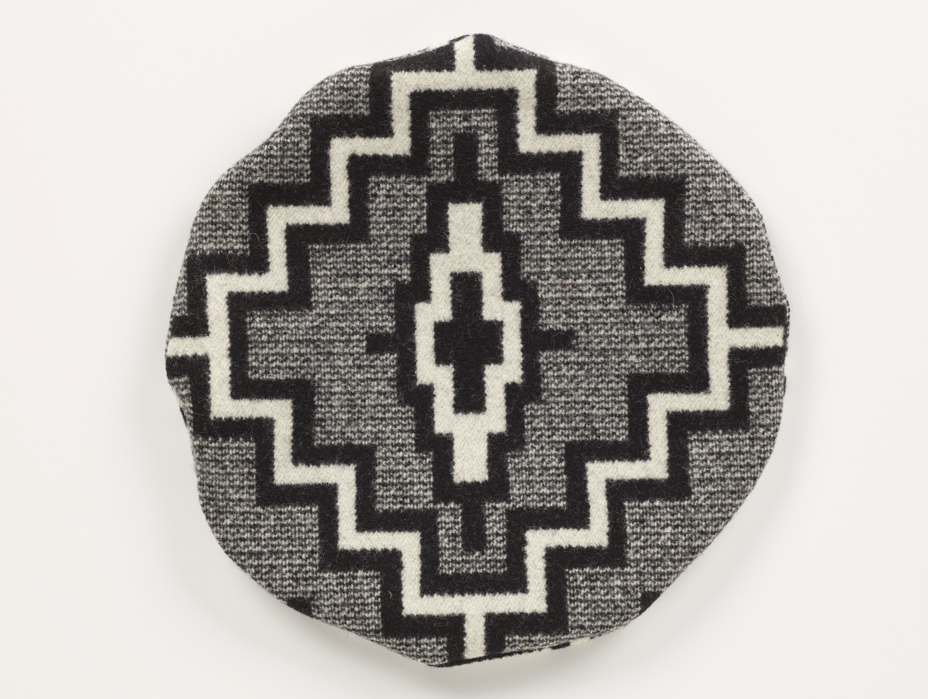 Top view of a gray tweed circular hat with a bold, black and white diamond-shaped center motif surrounded by a black-bordered white zig-zag design, displayed flat against a white background. 