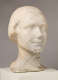 Three quarter front view of a blemished marble sculpture depicting the head of a female figure. The face features a faint smile and delicate details, framed by a short hairstyle.