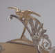 Detail of a golden bird with short beak and outstretched wings perched as if ready to fly from  silver wheat stalks decorating an ornate scroll. 