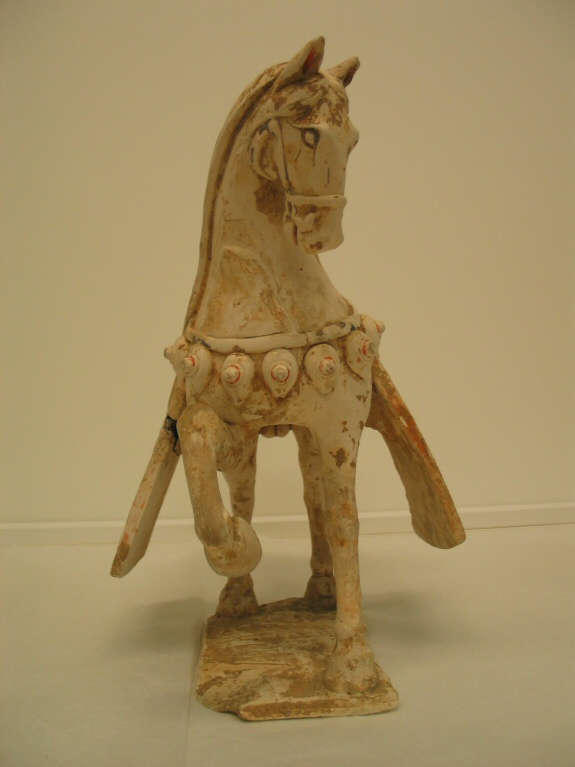 Front-view of a white ceramic sculpture of an ornamented saddled horse with one foot raised, looking aside, standing on a thin base. Belts and saddle blankets hang off its body.