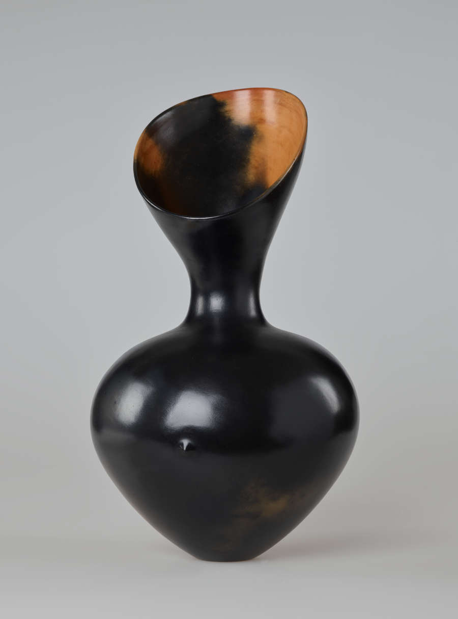 An asymmetrical, hourglass-shaped black vessel with a reflective finish. Its mouth slopes downward toward the viewer, exposing a bright terracotta interior.