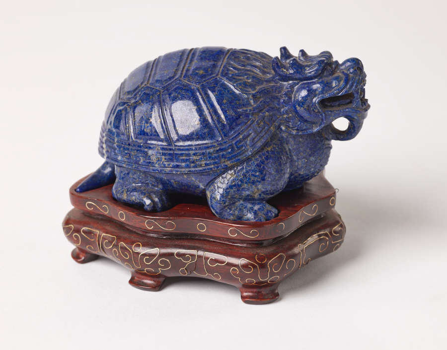 Side view of a blue sculpture of a horned tortoise, whose surface is covered with carved geometric patterns. It stands on a brown base with steps and golden patterning.