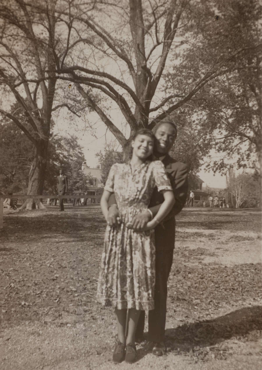 Vintage snapshot of a young dark-skinned man and light-skinned woman posing outside. They face the camera, smiling. He stands behind her with his hands at her waist. 
