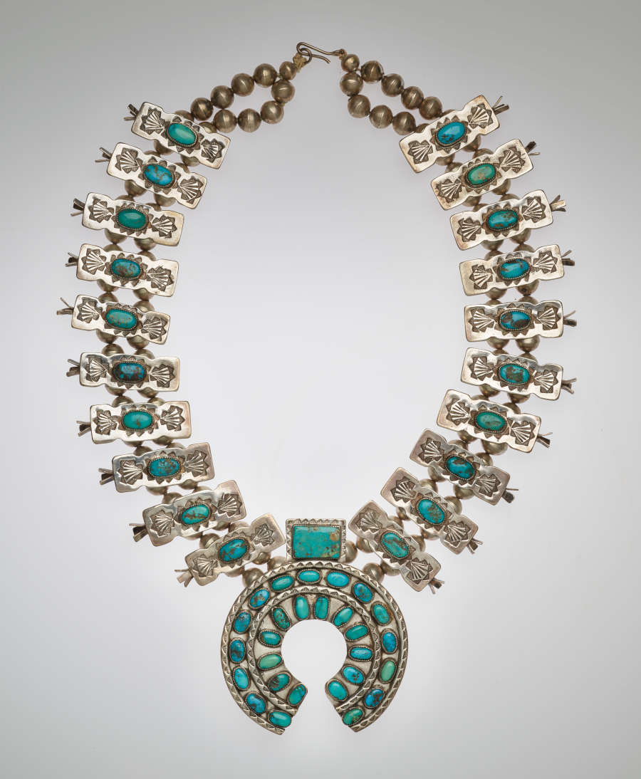 Silver and turquoise necklace. The chain features turquoise stones set within silver rectangular ornaments threaded into two rows of beaded chains. The central pendant is a turquoise stone-set horseshoe shape.