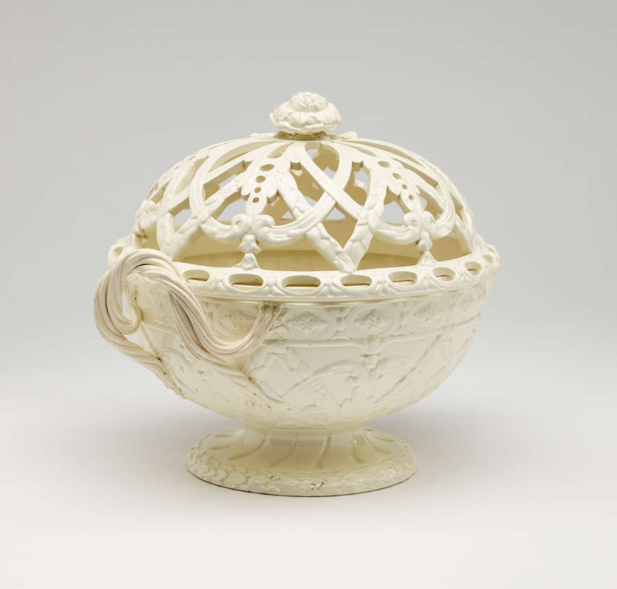 A cream-colored basket with sculptural woven handles, sculpted foot, and a lid with cutout designs.