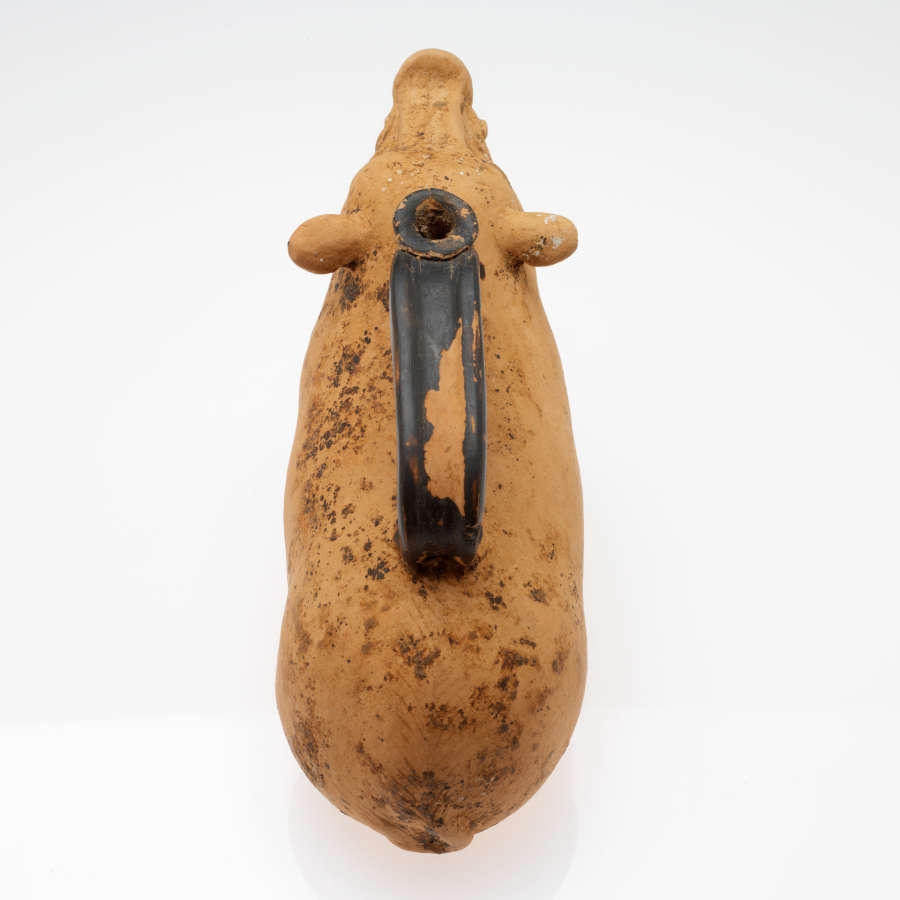 Top-view of a terracotta container in the form of a seated boar facing away from the viewer. Along its back is a long black handle with a narrow connected mouth.