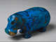 Bright blue ceramic hippopotamus figure decorated with black painted florals and reeds that adorn its short and rounded body. Patches of golden brown around the face indicate wear. 