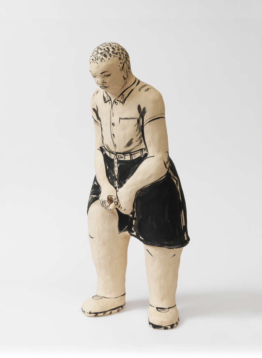 Stylized ceramic figure of a young boy leaning forward in a buttoned shirt tucked into dark shorts with his hands clasped in front of his pants, head tilted downward.
