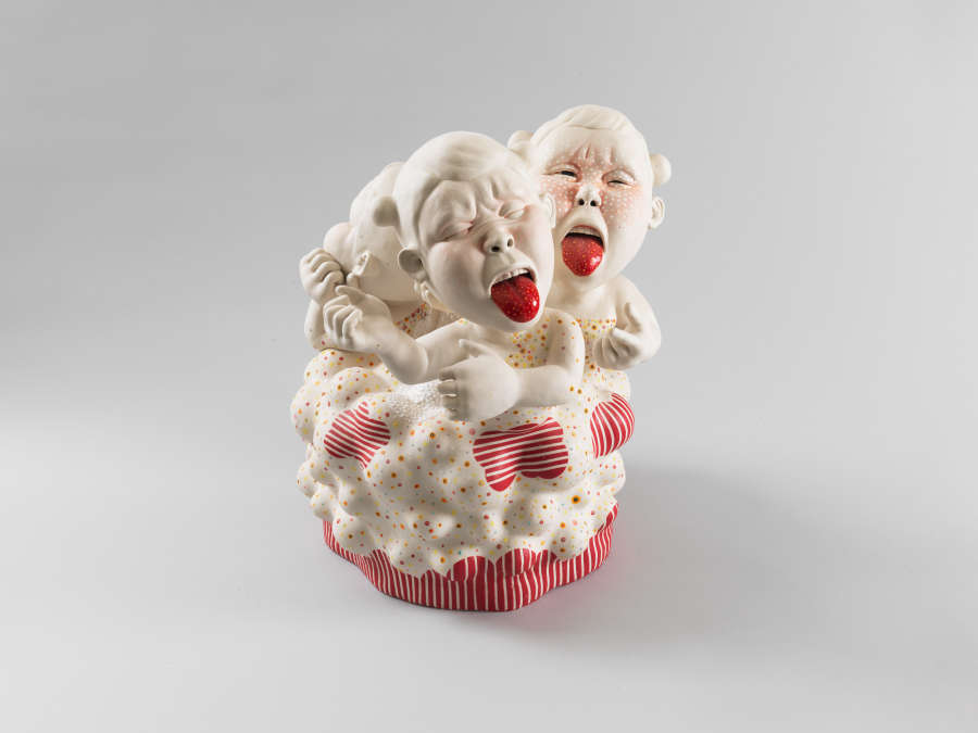 Sculpture of three childlike figures with oversized heads, bright red tongue sticking out of their exaggerated mouths, emerging from a circular, cupcake-like base decorated with thin red and white stripes.