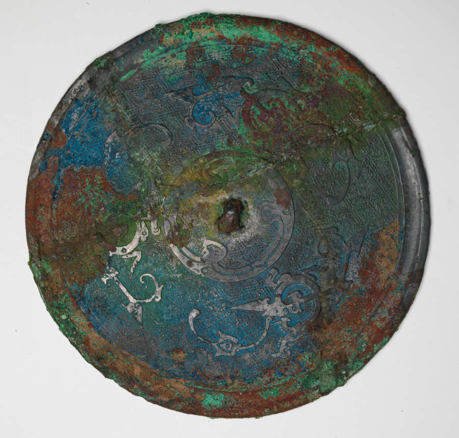 Back of an ornate circular mirror with raised patterning of organic, floral motifs arranged concentrically. Its worn surface is no longer reflective and instead consists of blues, greens, and browns.
