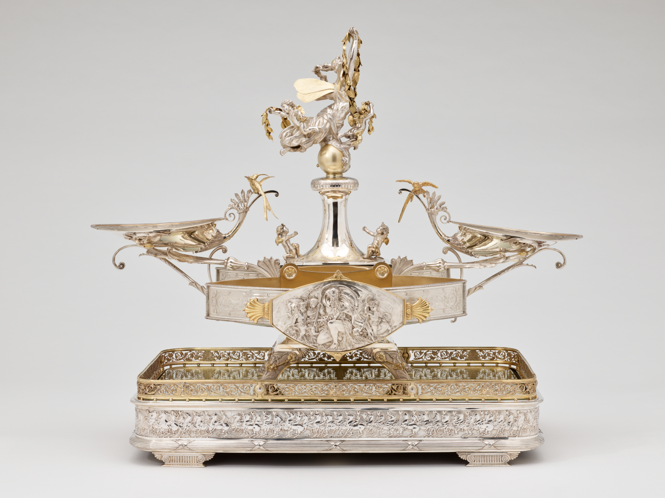 Profile view of elaborate silver double-oval centerpiece with swirling arms extending to two shallow side-dishes, with central fountain-like stem topped by classical figurines, all on a rectangular tray pedestal.  