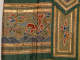 A mythological hoofed creature with red cloud-like wings surrounded by colorful clouds floating above blue waves, framed by horizontal gold bands, next to a long gold-embroidered green folded layer.
