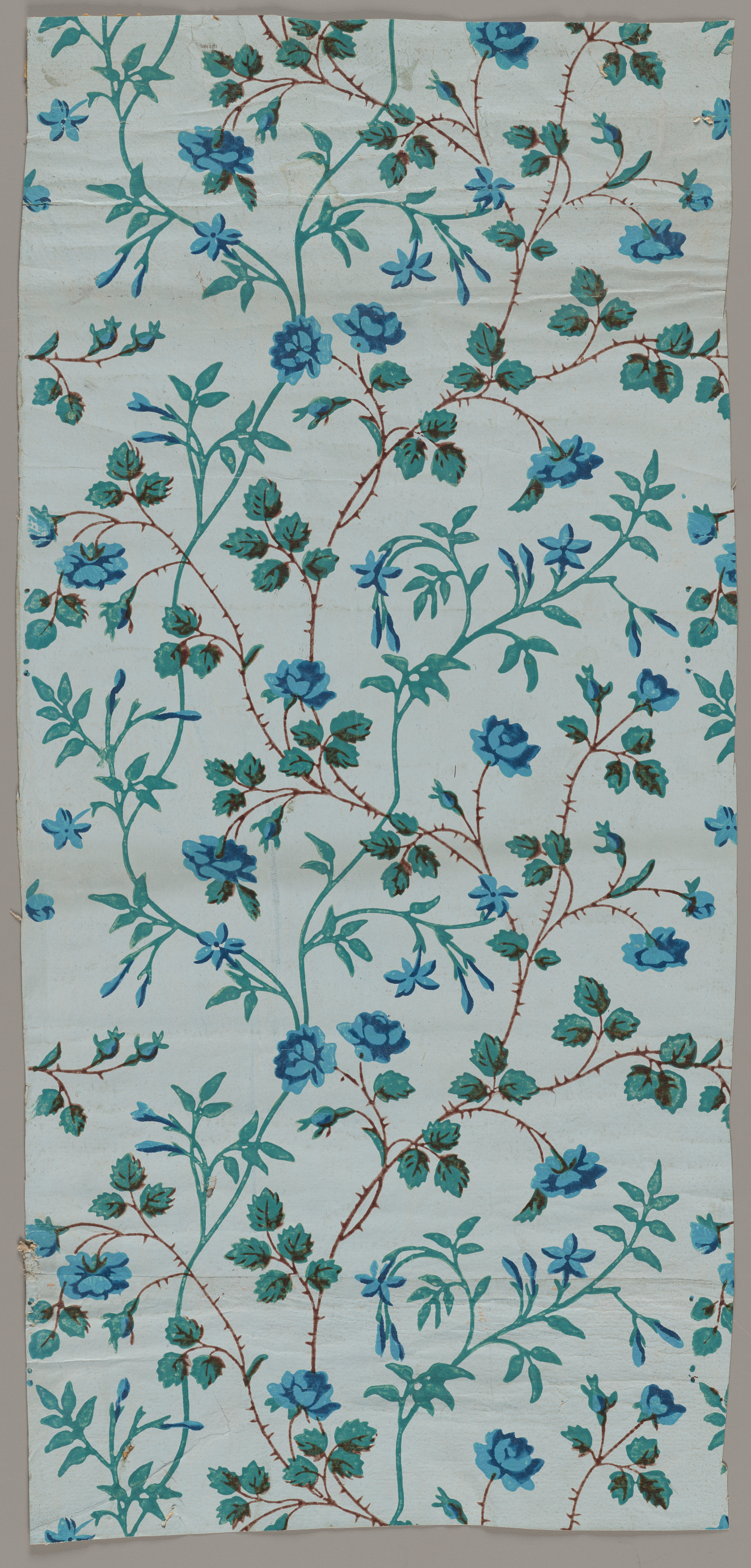 Light blue wallpaper of winding brown thorny branches being intersected by turquoise vines throughout. Multiple blue flowers are attached to the brown branches and turquoise leaves on the turquoise vines.