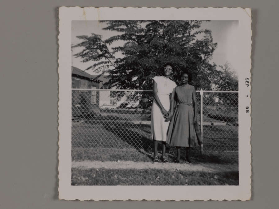 A snapshot of two young Black women smiling, holding hands, and leaning into each other. They stand by a fence in a yard.