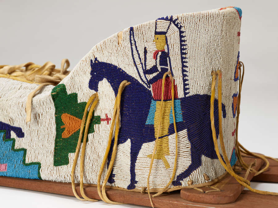 Detail of beaded object. White background beaded with blue horse and colorful rider in headdress. Some geometric decoration. Leather ties hanging down and lacing at top.