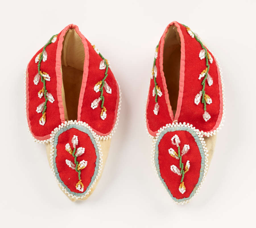 Top view of a pair of moccasins. Their tops each consist of two pieces of red fabric with green and white floral embroidery. The soles and interiors are tan.