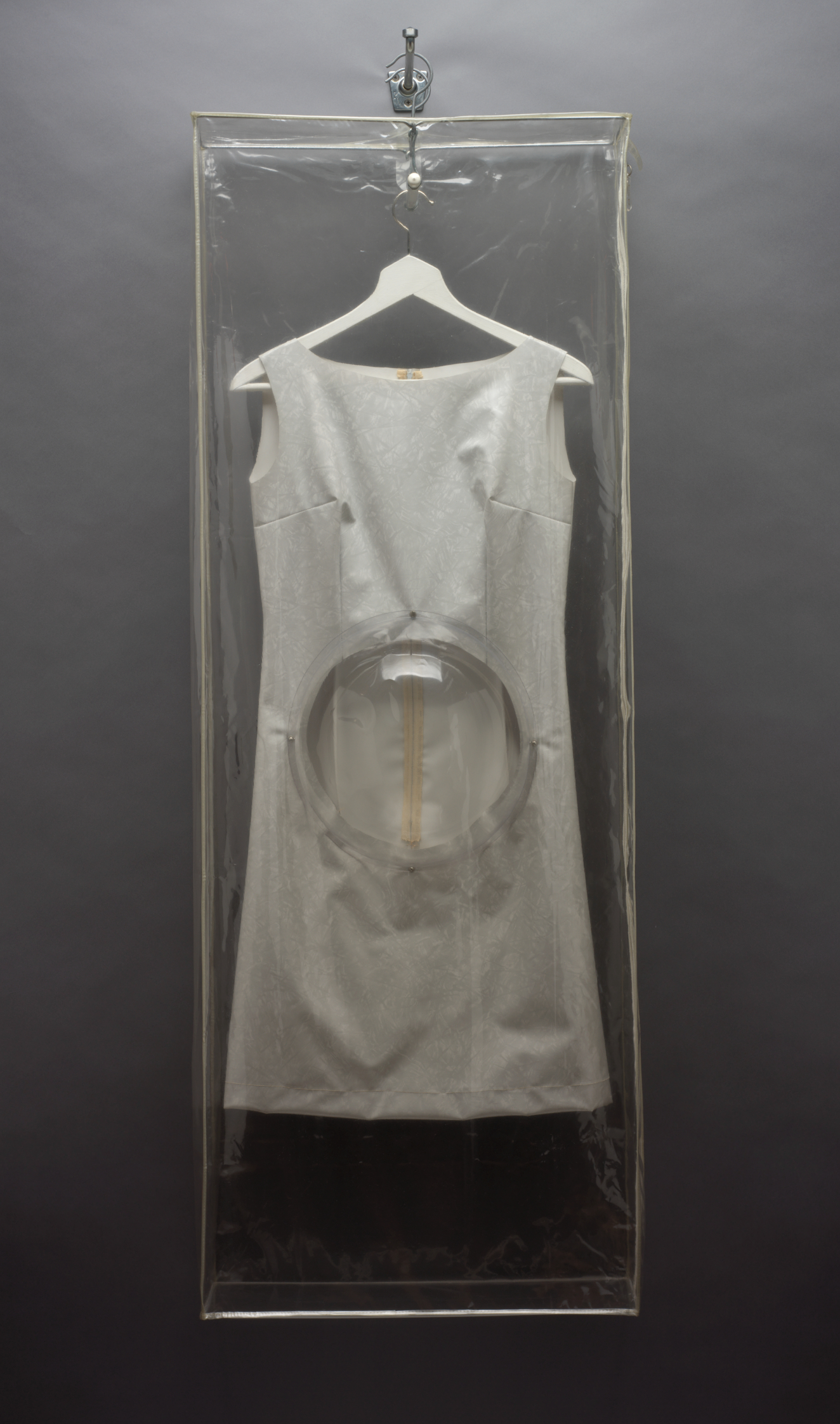 A light-colored sleeveless dress hanging in a clear garment bag featuring a transparent bubble-shaped panel around the abdomen area. 