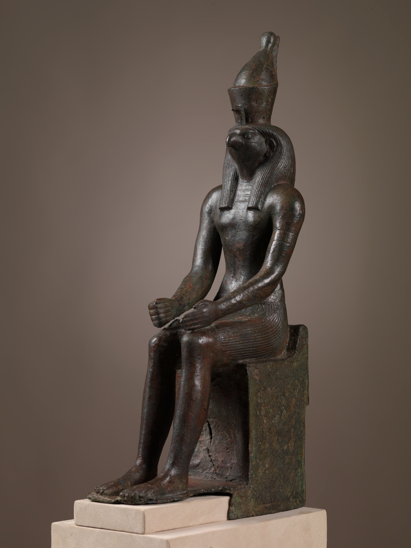 A seated statue of a human figure with a falcon head, muscular body, and tall headdress, seated on a rectangular throne.
