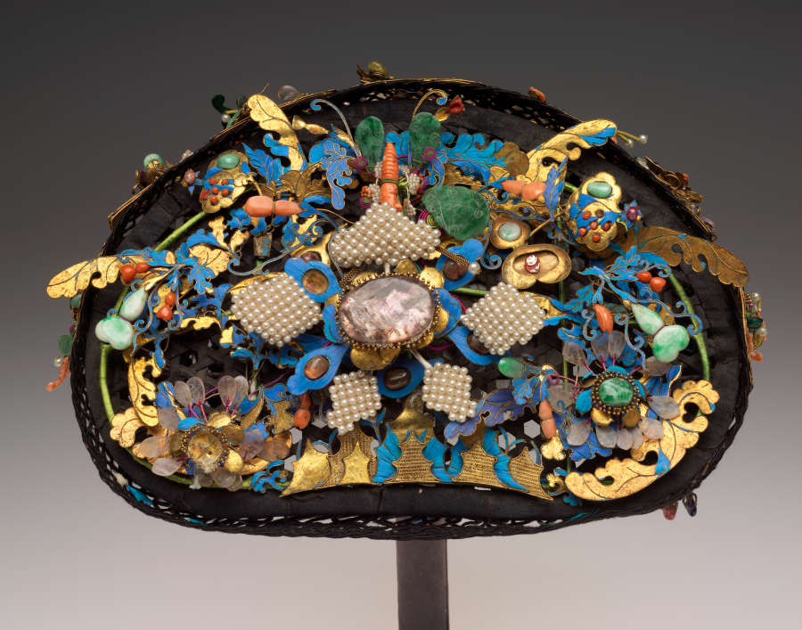 A headdress composed of several turquoise, golden, orange, green, and white stones, jewels, and feathers attached to a dark base, together taking the form of floral, organic motifs.