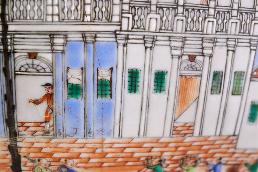 A close up of a painted ceramic showing a blue and white walled building with tall columns, green-paned windows, and tiled floors, featuring a figure stepping through a doorway.