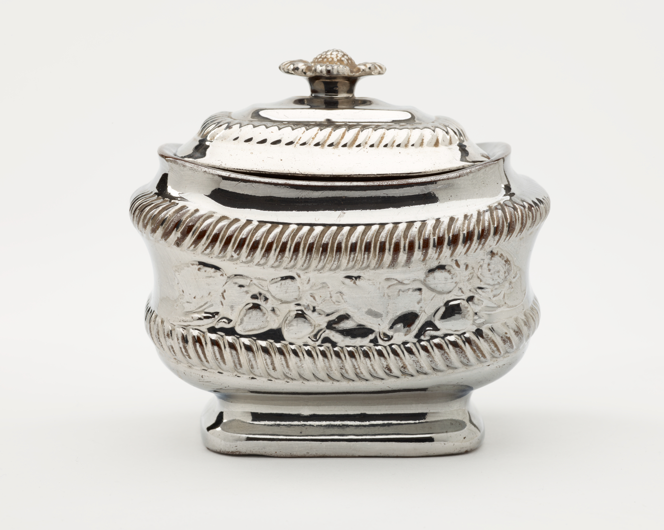 A silver sugar bowl with a decorative lid and a rounded square body with sculptural decorations.
