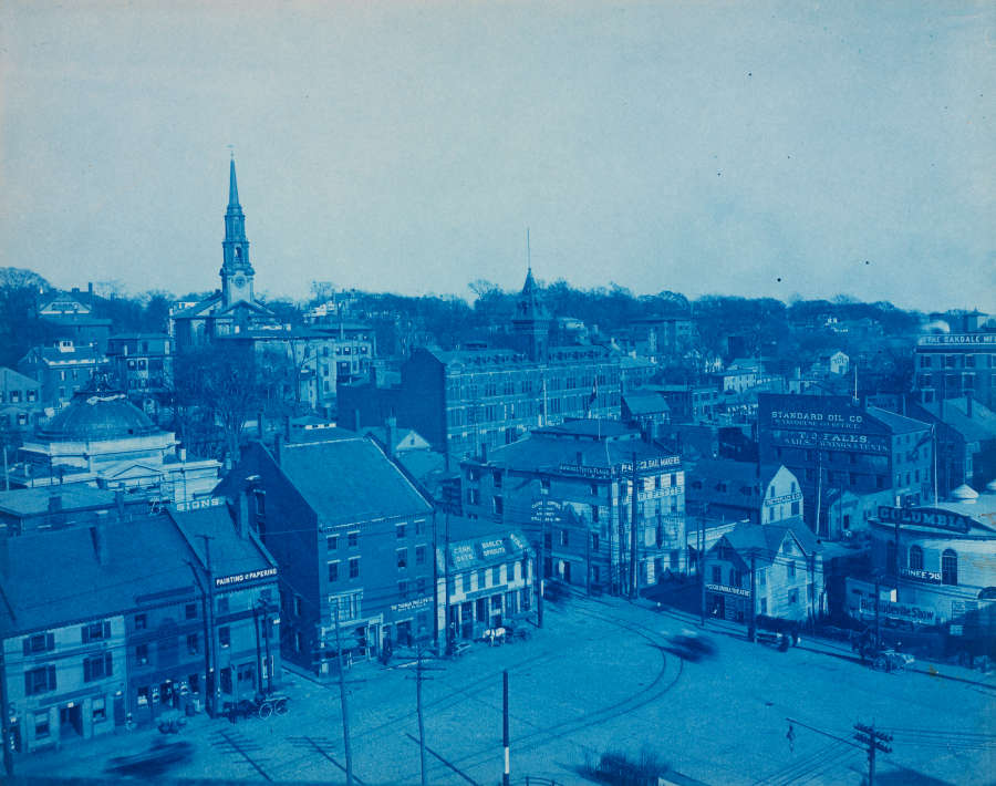 Antique blue-tinted aerial view of an old urban area. Historic buildings encircle the town square, which is crisscrossed by tire tracks. A steepled church oversees the scene.