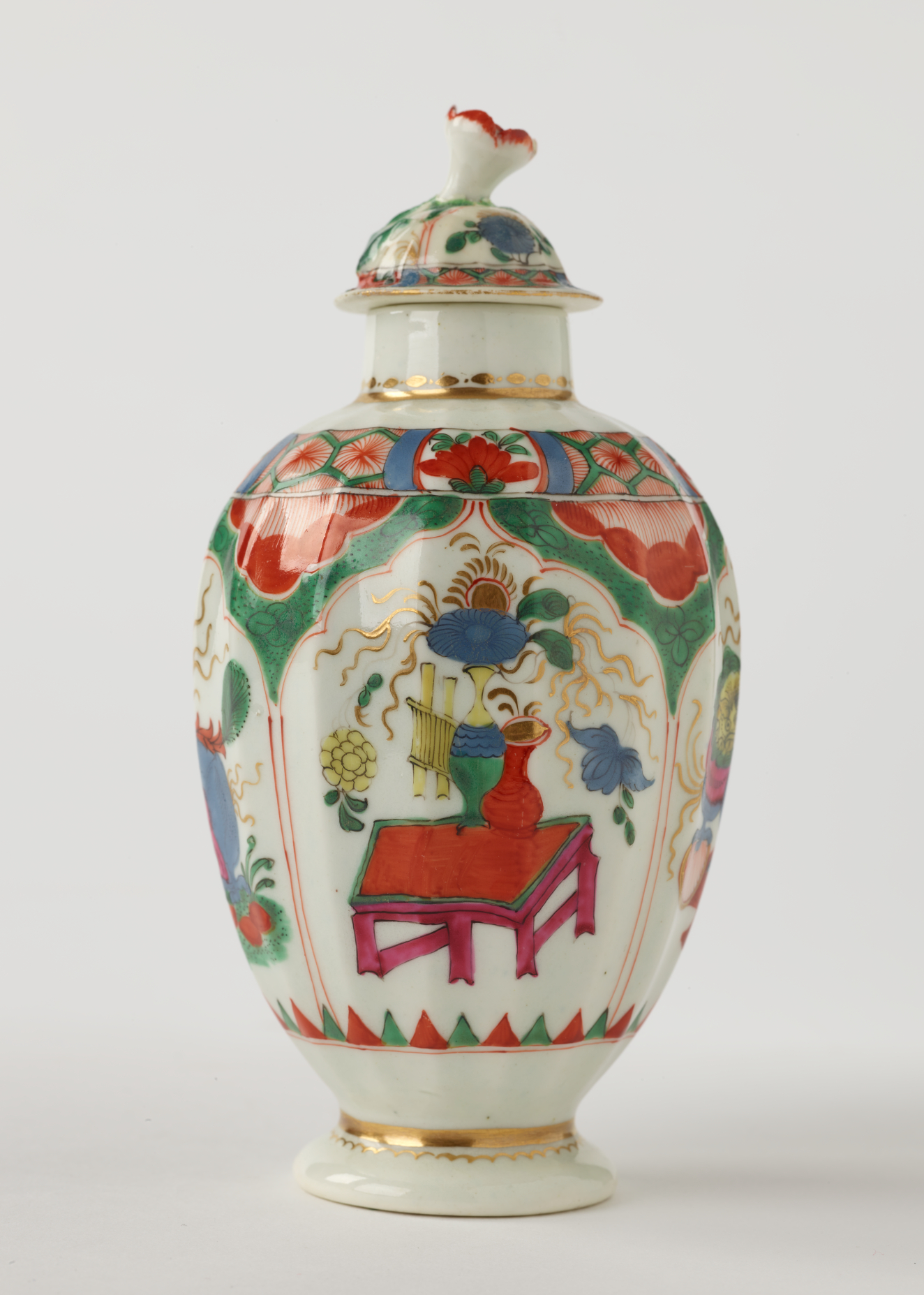 A tea caddy with a lid and green, red, blue, pink, yellow and gilded decorations. The finial is a sculptural flower white, red and green.