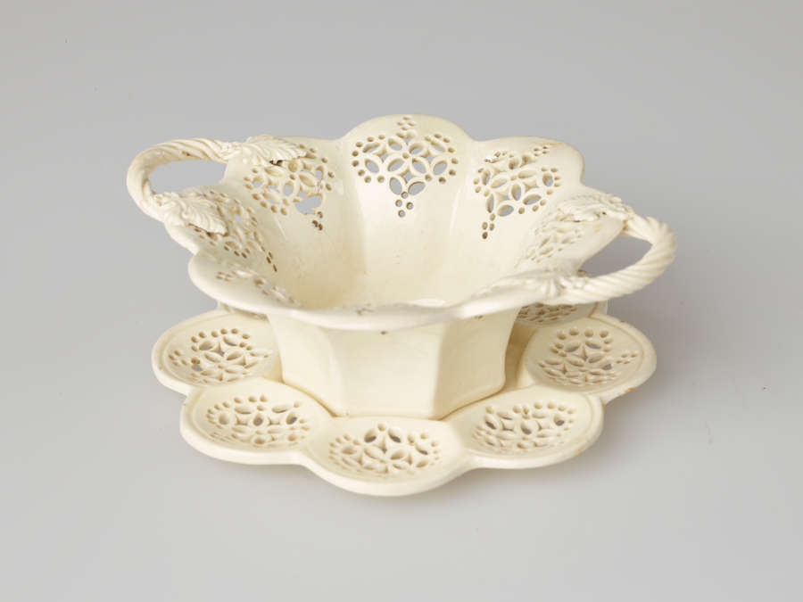 A cream-colored dish with two swirled handles and pierced decorations. Dish sits on a matching tray.