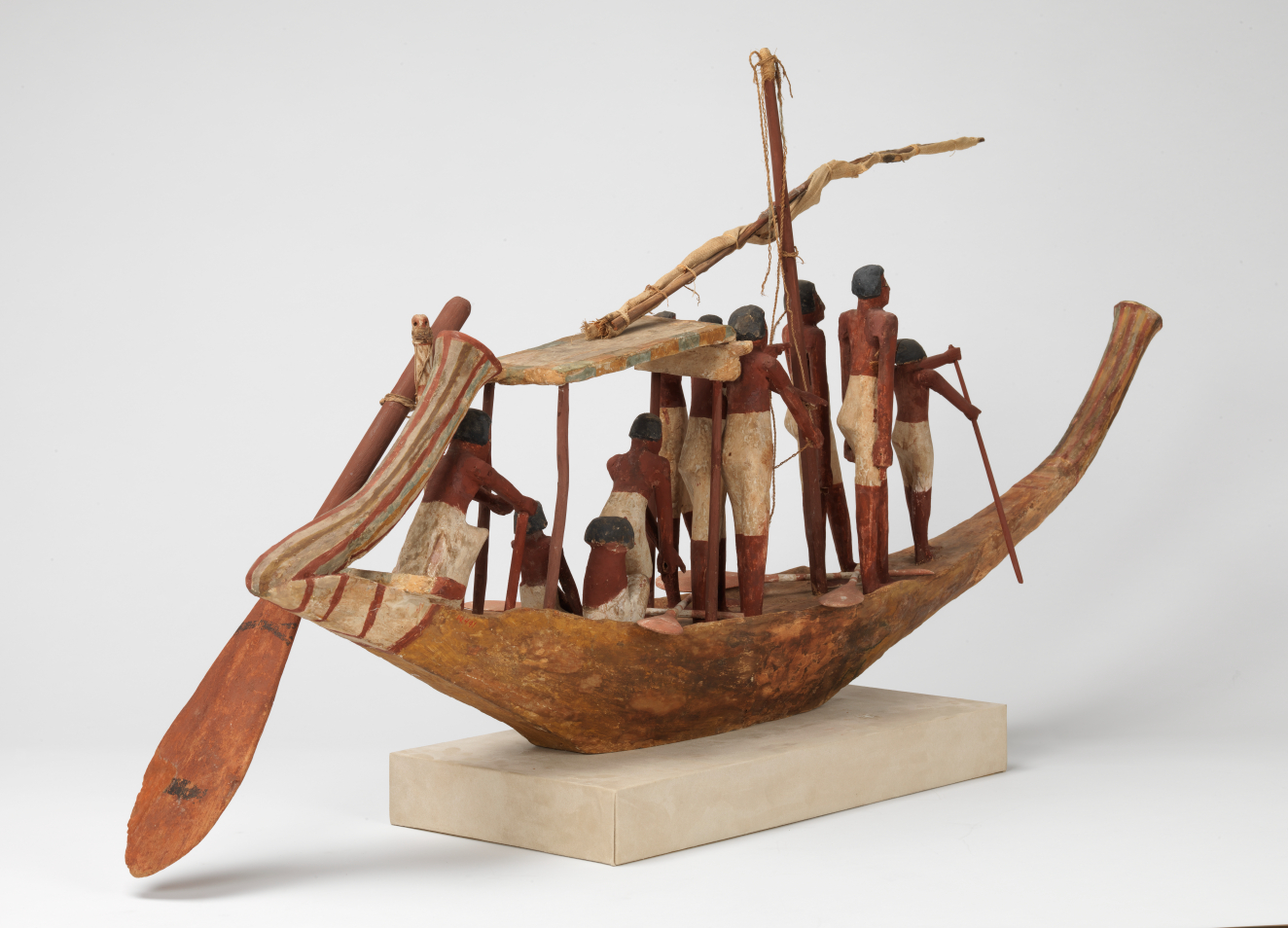 A carved wooden boat model on a rectangular off-white base with a tied up sail manned by sailors with brown skin and white trousers holding oars and ropes.