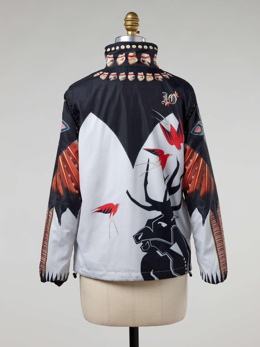 Back of a windbreaker on a mannequin. Its top features a black angular design with red-orange feather and shell-motifs. Its white bottom features the silhouette of an antlered animal.